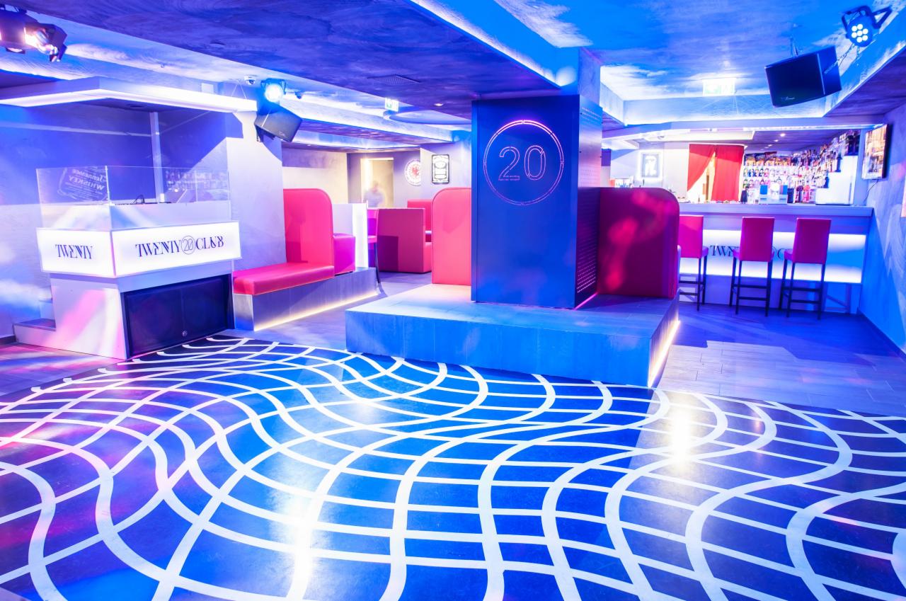 Nowoczesne wnętrze klubu nocnego z geometrycznym wzorem na podłodze, oświetleniem LED w odcieniach niebieskiego i różu, oraz barem w tle.