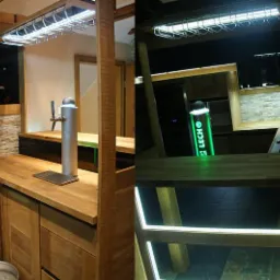 Drewniany bar z blatem z jasnego drewna, chromowanym kranem do piwa i podświetlanym logo 'LECH', oświetlony od góry lampą z uchwytami na kieliszki.