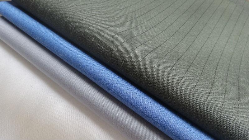 Stos trzech próbek tkanin: ciemnoszary w prążki, błękitny i jasnoszary, ułożonych warstwami, ukazujących teksturę materiałów.