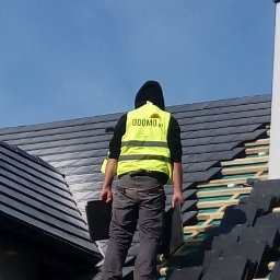 Odomo.pl Sp. z o.o. - Pracownik w kamizelce z logo firmy na dachu krytym czarną dachówką, w trakcie układania. Widoczne elementy konstrukcji dachu i komin. Błękitne niebo w tle.