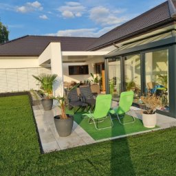 Odomo.pl Sp. z o.o. - Nowoczesny dom z przeszkloną oranżerią i tarasem z zielonymi krzesłami, donicami z palmami i zadbanym trawnikiem w słoneczny dzień.