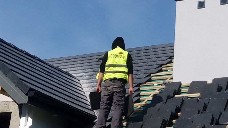 Pracownik w kamizelce z logo firmy na dachu krytym czarną dachówką, w trakcie układania. Widoczne elementy konstrukcji dachu i komin. Błękitne niebo w tle.