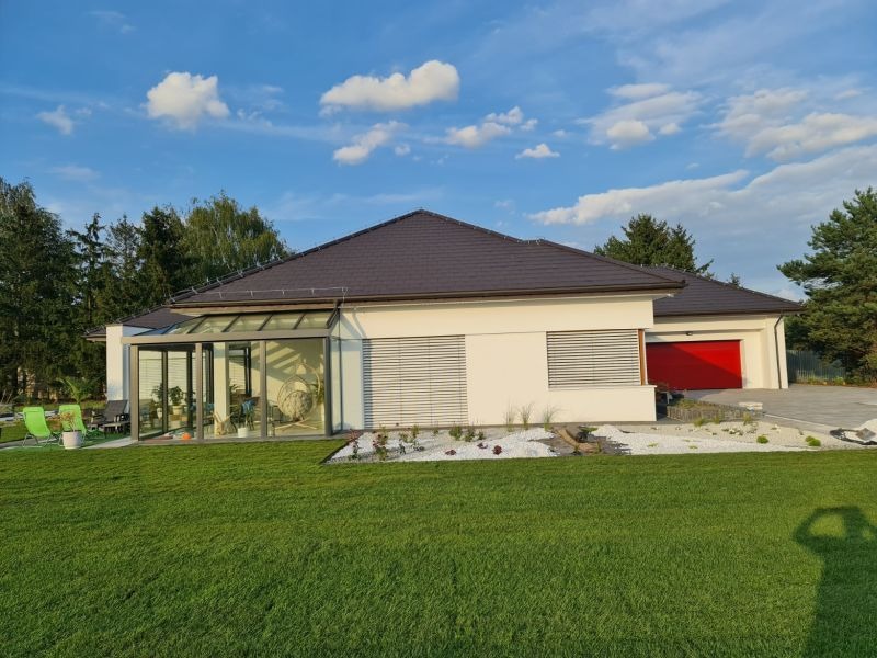 Nowoczesny dom z przeszkloną oranżerią i czerwonym garażem, otoczony zielonym trawnikiem i dekoracyjnym ogrodem z białym żwirem. Błękitne niebo z chmurami w tle.