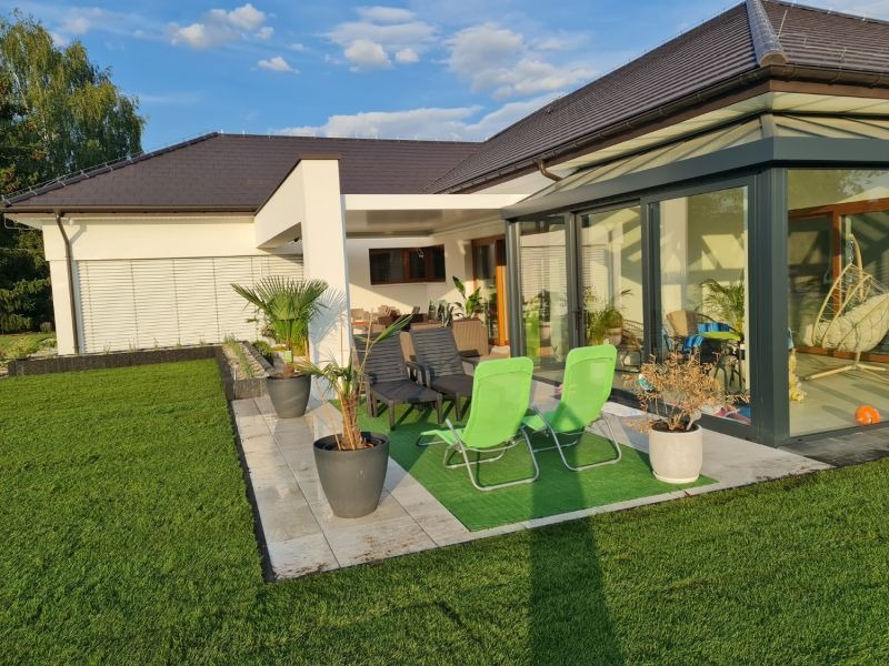 Nowoczesny dom z przeszkloną oranżerią i tarasem z zielonymi krzesłami, donicami z palmami i zadbanym trawnikiem w słoneczny dzień.