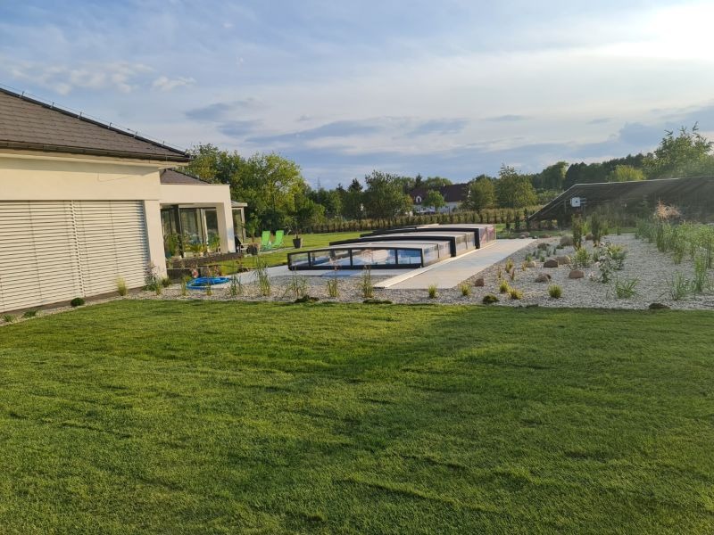 Nowoczesny dom z basenem i zadaszeniem, otoczony trawnikiem i ozdobnym ogrodem z białym żwirem. Widok na budynek z roletami zewnętrznymi i zielone otoczenie.