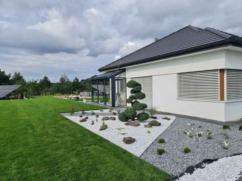 Nowoczesny dom z ogrodem: biała elewacja, szare rolety, przeszklona weranda, trawnik i dekoracyjny ogródek z białym i szarym żwirem oraz formowanymi krzewami.