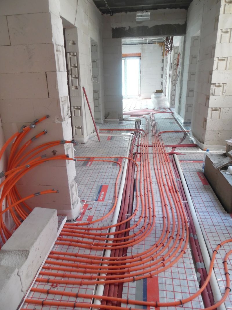 Instalacja ogrzewania podłogowego z pomarańczowymi rurami na izolacji, w surowym wnętrzu budynku z białych bloczków. Widok na korytarz z otworami drzwiowymi.
