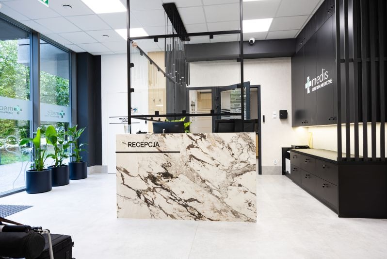 Nowoczesne wnętrze recepcji w budynku komercyjnym z marmurowym blatem, czarnymi szafkami i roślinami w donicach. Widoczne logo 'medis centrum medyczne'.