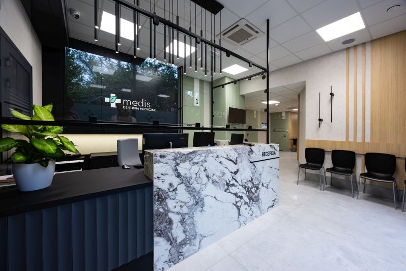 Nowoczesne wnętrze recepcji centrum medycznego z marmurowym blatem, czarnymi krzesłami i rośliną doniczkową. Widoczne logo centrum na szklanej ścianie.
