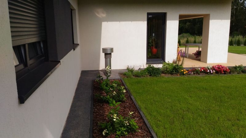 Elewacja domu z szarymi oknami i wentylacją, rabata kwiatowa z korą, trawnik. W tle widoczny taras i zieleń. Ujęcie z poziomu gruntu, w słoneczny dzień.