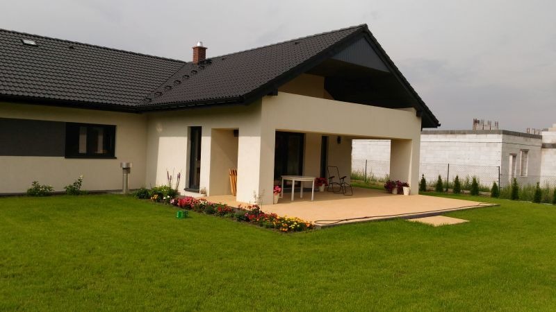Parterowy dom z ciemnym dachem i jasną elewacją, zadaszony taras z meblami ogrodowymi, rabata kwiatowa i trawnik. W tle budynek w trakcie budowy.