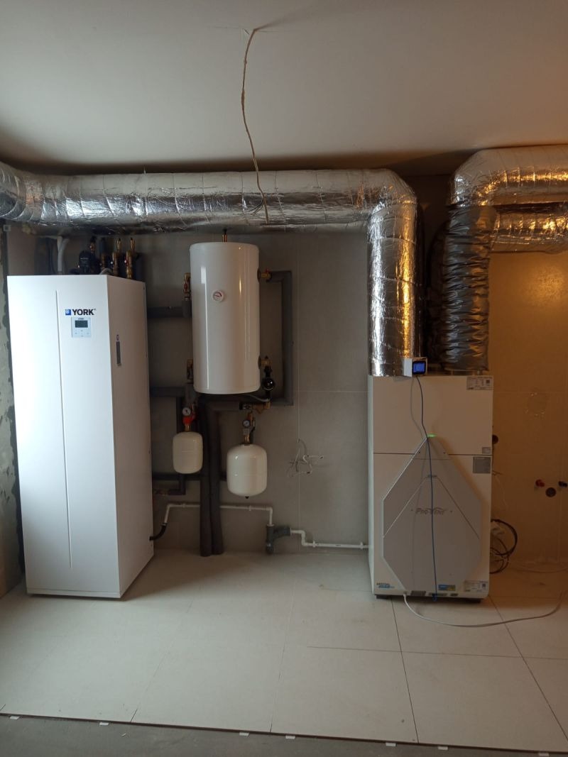 Biała pompa ciepła York i rekuperator z srebrnymi rurami wentylacyjnymi w pomieszczeniu z szarą ścianą i jasną podłogą.