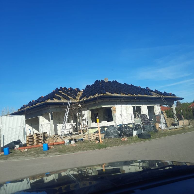 Budowa domu jednorodzinnego w trakcie układania dachówki, widoczne rusztowania i materiały budowlane. Stan surowy otwarty, błękitne niebo w tle.