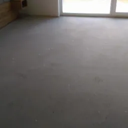 Szara, gładka powierzchnia wylewki w nowym pomieszczeniu z widoczną ramą okienną i elementami wykończenia ściany.