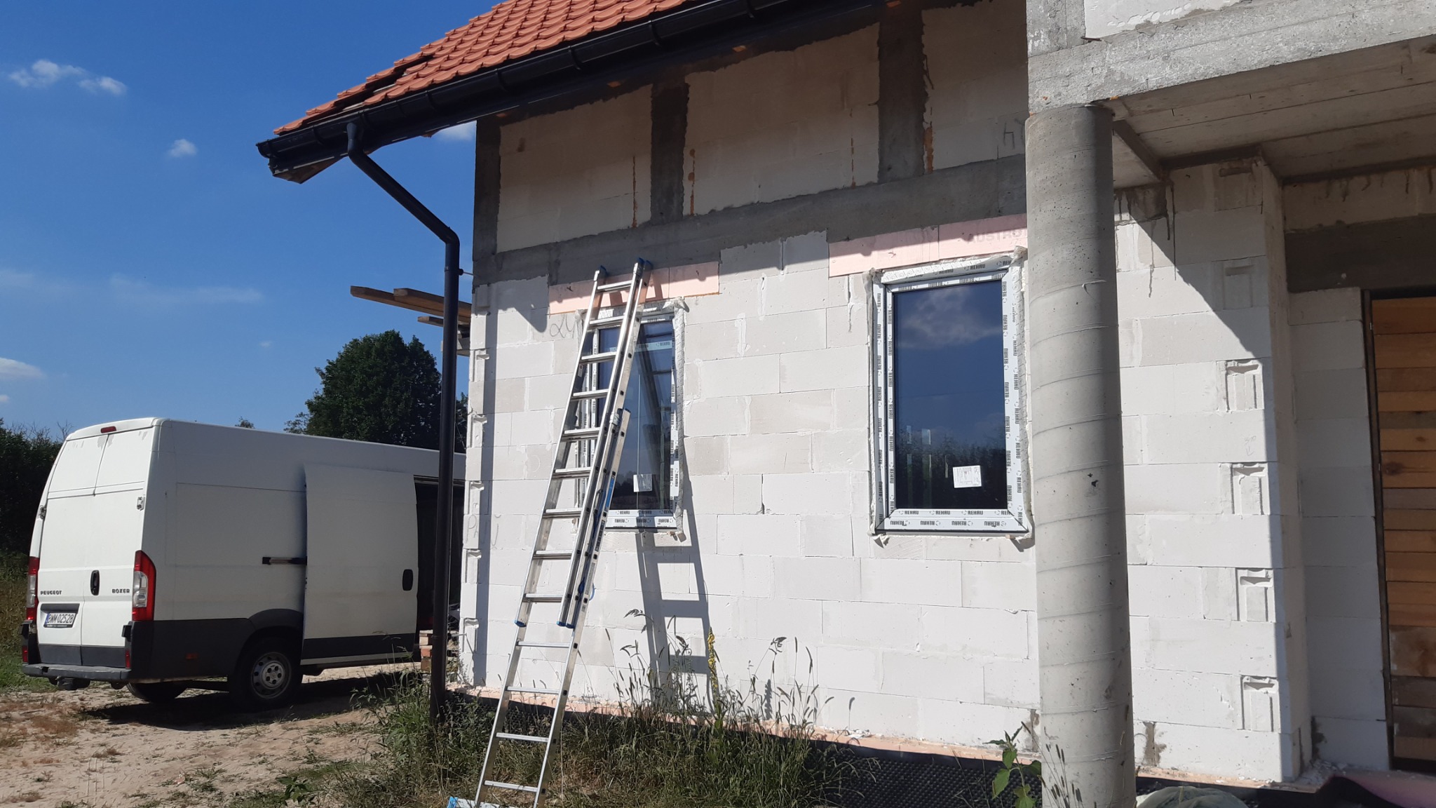 Montaż okien w nowo budowanym domu w Czyżewie; widoczna drabina oparta o ścianę z zamontowanym oknem, biały bus dostawczy w tle.