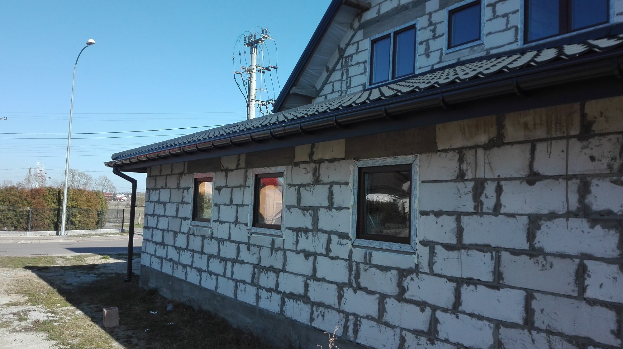 Surowy budynek z białego bloczka z zamontowanymi oknami z brązowymi ramami, widoczne profile montażowe, w tle słup elektryczny i latarnia uliczna.