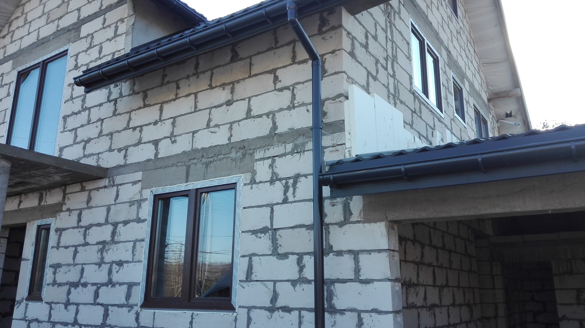 Surowy stan budynku w trakcie budowy z zamontowanymi oknami z brązowymi ramami i orynnowaniem w kolorze grafitowym.