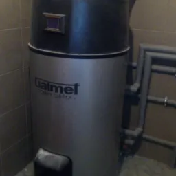 Szara pompa ciepła Galmet z czarną górną częścią i wyświetlaczem, widoczne rury wentylacyjne i instalacyjne w pomieszczeniu z szarą glazurą.