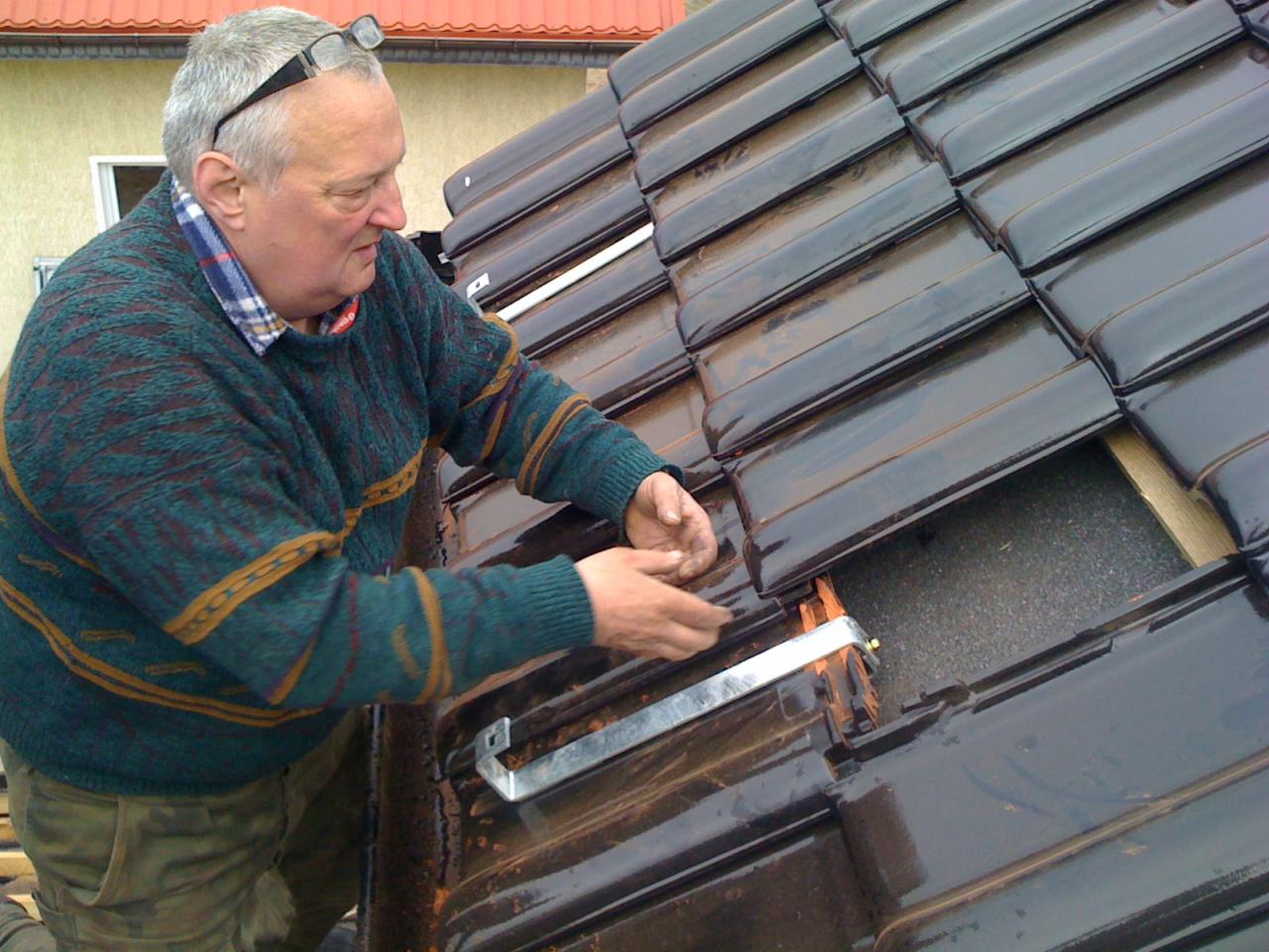 Monter w swetrze mocuje metalowy element do ciemnej, błyszczącej dachówki na skośnym dachu, widoczna izolacja pod dachówką.