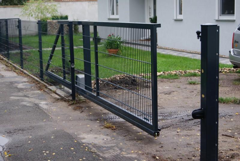 Automatyczna brama przesuwna z metalową siatką, częściowo otwarta, z widocznym napędem i słupkiem sterującym, na tle domu z trawnikiem