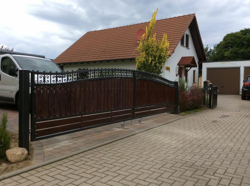 Dwuskrzydłowa brama wjazdowa z drewnianym wypełnieniem i kutymi elementami dekoracyjnymi, zamontowana na posesji z domem jednorodzinnym i garażem.