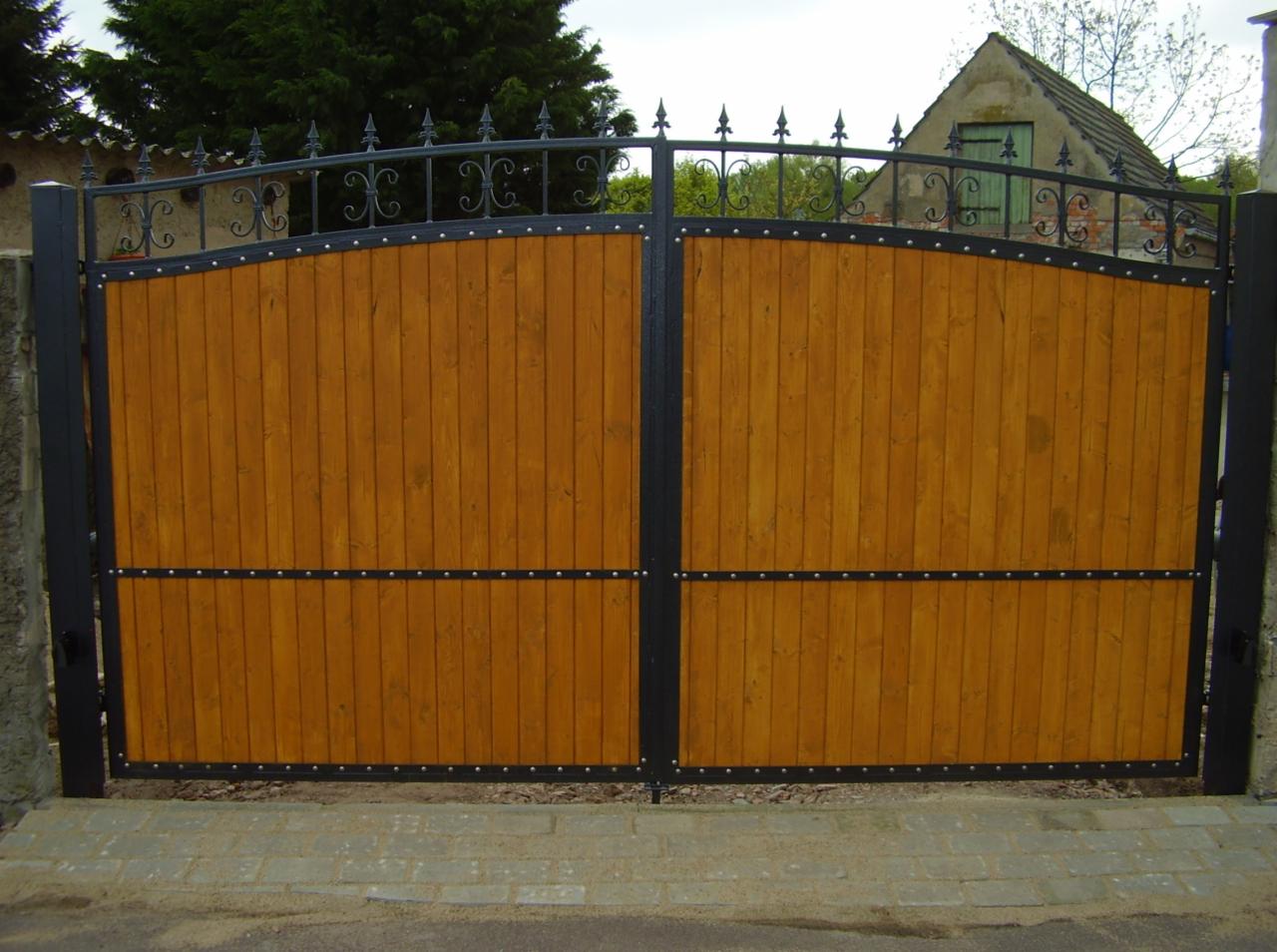 Dwuskrzydłowa brama wjazdowa z pionowych desek w kolorze miodowego drewna, z czarną metalową ramą i ozdobnym kutym zwieńczeniem, zamontowana na betonowych słupkach.