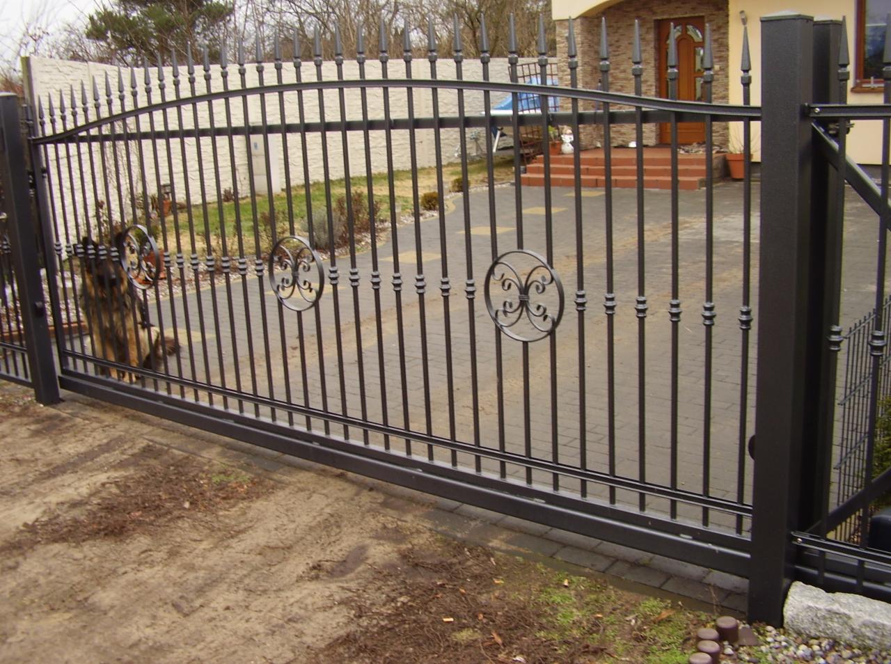 Metalowa brama przesuwna z dekoracyjnymi rozetami i szpikulcami, widok z zewnątrz posesji, częściowo widoczny pies za bramą.
