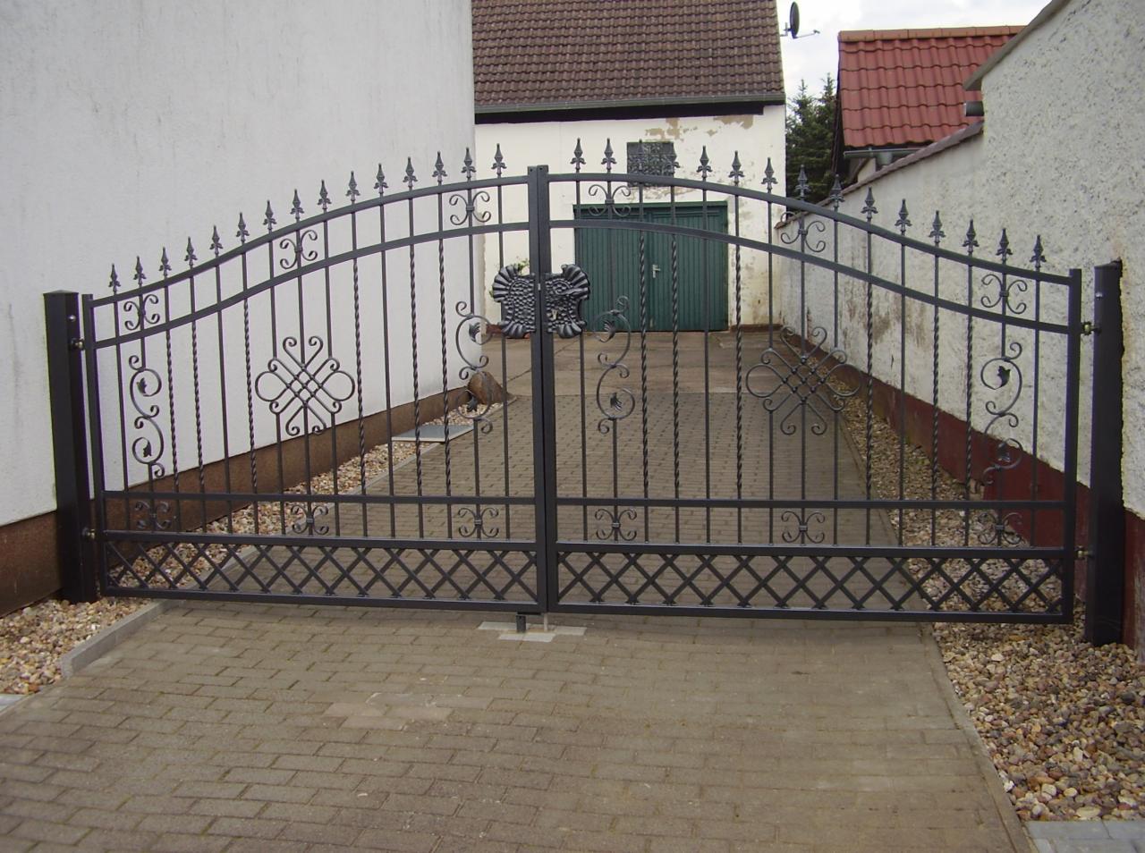 Dwuskrzydłowa brama kuta z dekoracyjnymi elementami, prowadząca na wybrukowany podjazd, widoczna z przodu na tle białych ścian i zielonych drzwi garażowych.