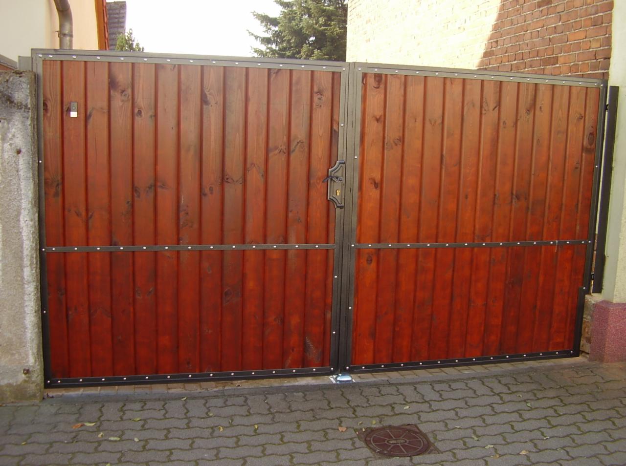 Dwuskrzydłowa brama wjazdowa z pionowych desek o ciepłym, brązowym odcieniu, obramowana czarną, metalową ramą z widocznymi nitami, zamknięta na zamek z ozdobną rękojeścią. Brama osadzona...