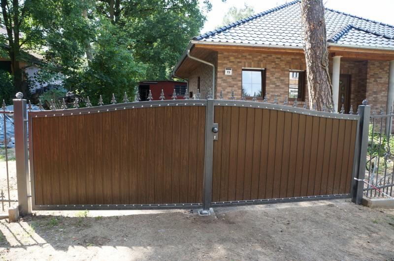 Dwuskrzydłowa brama wjazdowa z wypełnieniem imitującym drewno, stalowa rama i ozdobne groty na górze, widok na dom z cegły w tle.