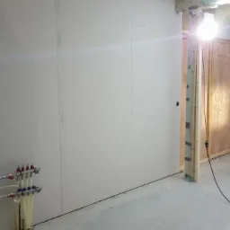 Montaż płyt gipsowo-kartonowych na ścianie podczas remontu z widocznymi rurami instalacji grzewczej i oświetleniem budowlanym.