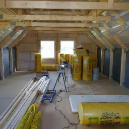 Wnętrze poddasza w trakcie izolacji, widoczne belki konstrukcyjne, folia paroizolacyjna, materiały izolacyjne Isover w rolkach, narzędzia pracy i przygotowane deski.