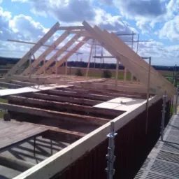 Konstrukcja drewniana dachu w trakcie budowy, widoczne krokwie i deski szalunkowe, w tle budynek z wieżą i pola uprawne pod błękitnym niebem.