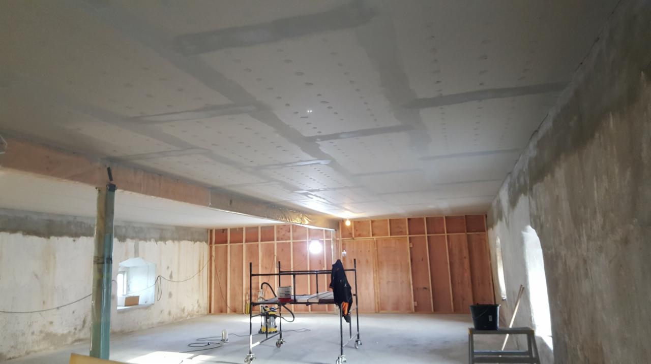 Wnętrze w trakcie prac wykończeniowych: ściany częściowo otynkowane, drewniana konstrukcja ścian działowych, sufit po szpachlowaniu, widoczne rusztowanie i narzędzia.