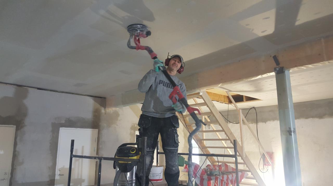 Pracownik na rusztowaniu szlifuje sufit gipsowy za pomocą szlifierki teleskopowej z odkurzaczem, w tle widoczne schody drewniane i surowe ściany.