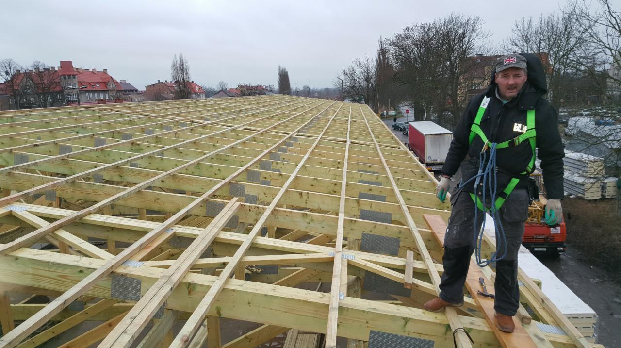 Pracownik w uprzęży zabezpieczającej stoi na drewnianej konstrukcji dachu domu w trakcie budowy, widoczne elementy wiązarów i łączników, w tle zabudowa miejska i pochmurne niebo.
