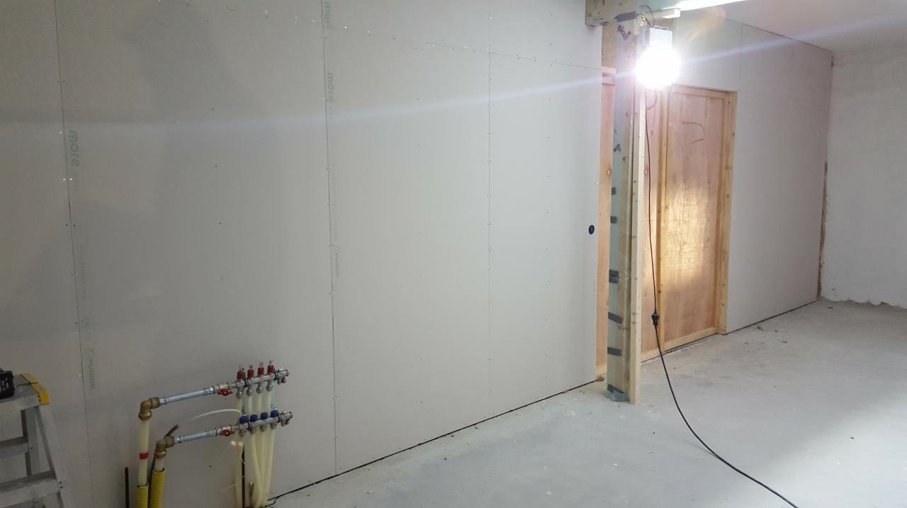 Montaż płyt gipsowo-kartonowych na ścianie podczas remontu z widocznymi rurami instalacji grzewczej i oświetleniem budowlanym.