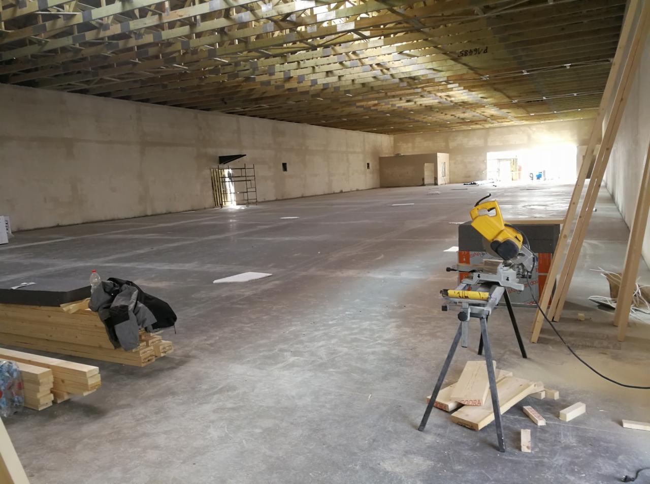Wnętrze hali w trakcie budowy, widoczny drewniany szkielet dachu, surowe ściany, betonowa posadzka, piła stołowa i przygotowane drewniane elementy.