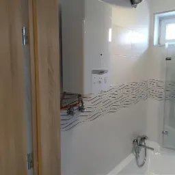 Biały piec gazowy Vaillant zamontowany na ścianie z dekoracyjnymi płytkami w łazience z częściowo widoczną, drewnianą ościeżnicą drzwiową i kabiną prysznicową.