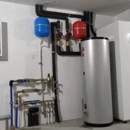 Zestaw urządzeń w kotłowni: biała pompa ciepła obok srebrnego zasobnika ciepłej wody, instalacja hydrauliczna z czarnymi rurami izolowanymi, czerwony i niebieski zbiornik wyrównawczy oraz skrzynka...