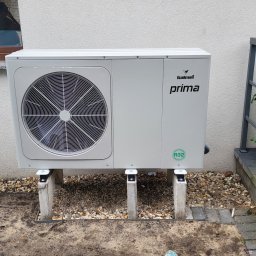 ASM eko-tech Adam Smolarczyk - Biała pompa ciepła Galmet Prima, zamontowana na betonowych wspornikach przy jasnej ścianie budynku, obok roślin w donicach i szarej balustrady.