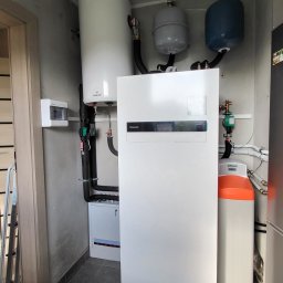 ASM eko-tech Adam Smolarczyk - Biała pompa ciepła Panasonic w kotłowni, z widocznym zasobnikiem Galmet, zbiornikami wyrównawczymi i rozdzielnicą elektryczną. Szare płytki podłogowe.