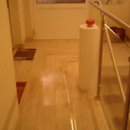 Polerowana marmurowa posadzka z delikatnym wzorem, fragment ściany, białe drzwi w tle, schody z metalową balustradą i szklanymi panelami, na słupku dekoracyjnym leży czerwone jabłko.