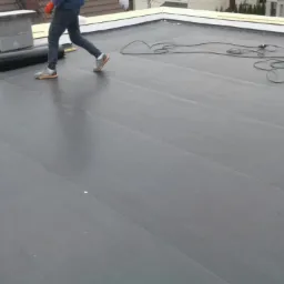 Czarny, płaski dach pokryty papą termozgrzewalną z widocznym kominem, narzędziami dekarskimi i fragmentem sylwetki dekarza w pomarańczowych rękawicach.