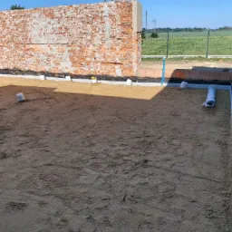 Przygotowany teren pod wylewkę fundamentów z widoczną izolacją przeciwwilgociową i częściowo wymurowaną ścianą z cegły, na ziemi widoczne ślady stóp, w tle ogrodzenie i pole.