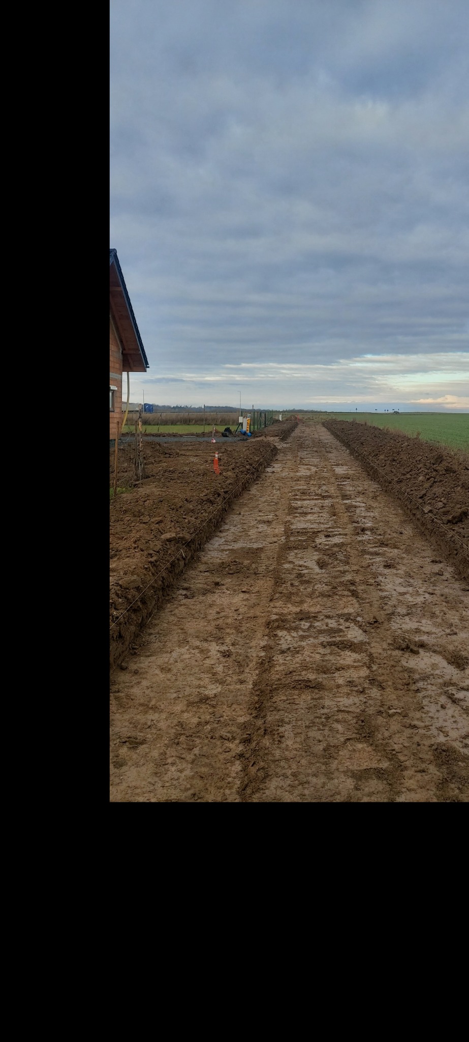 Przygotowanie terenu pod budowę domu z wykopanymi fundamentami, widoczne ślady opon ciężkiego sprzętu, w tle częściowo wykończony budynek w stanie surowym otwartym i zielone pole.