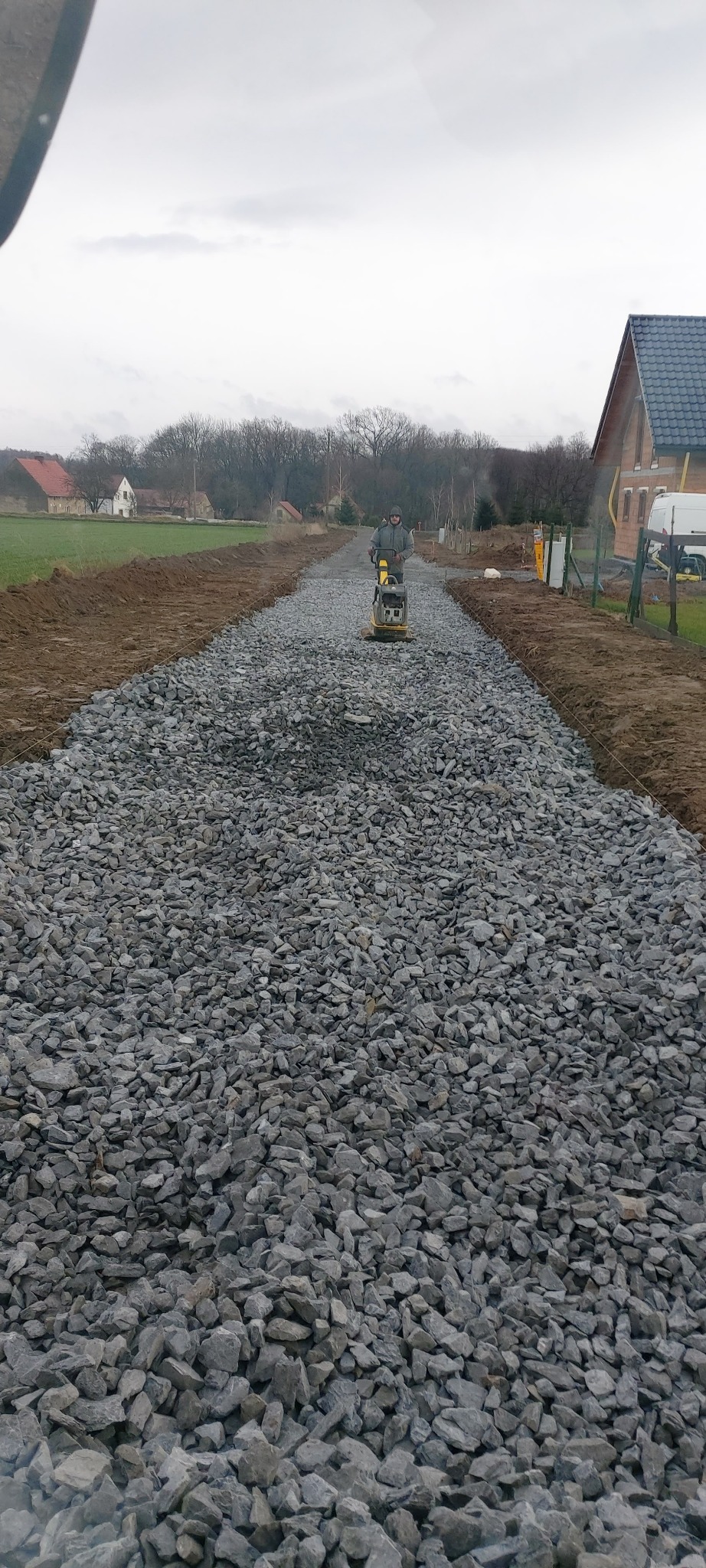 Pracownik zagęszcza kamienny podkład pod drogę dojazdową za pomocą zagęszczarki wibracyjnej, widoczny nowy dom jednorodzinny w tle i przygotowane obrzeża.