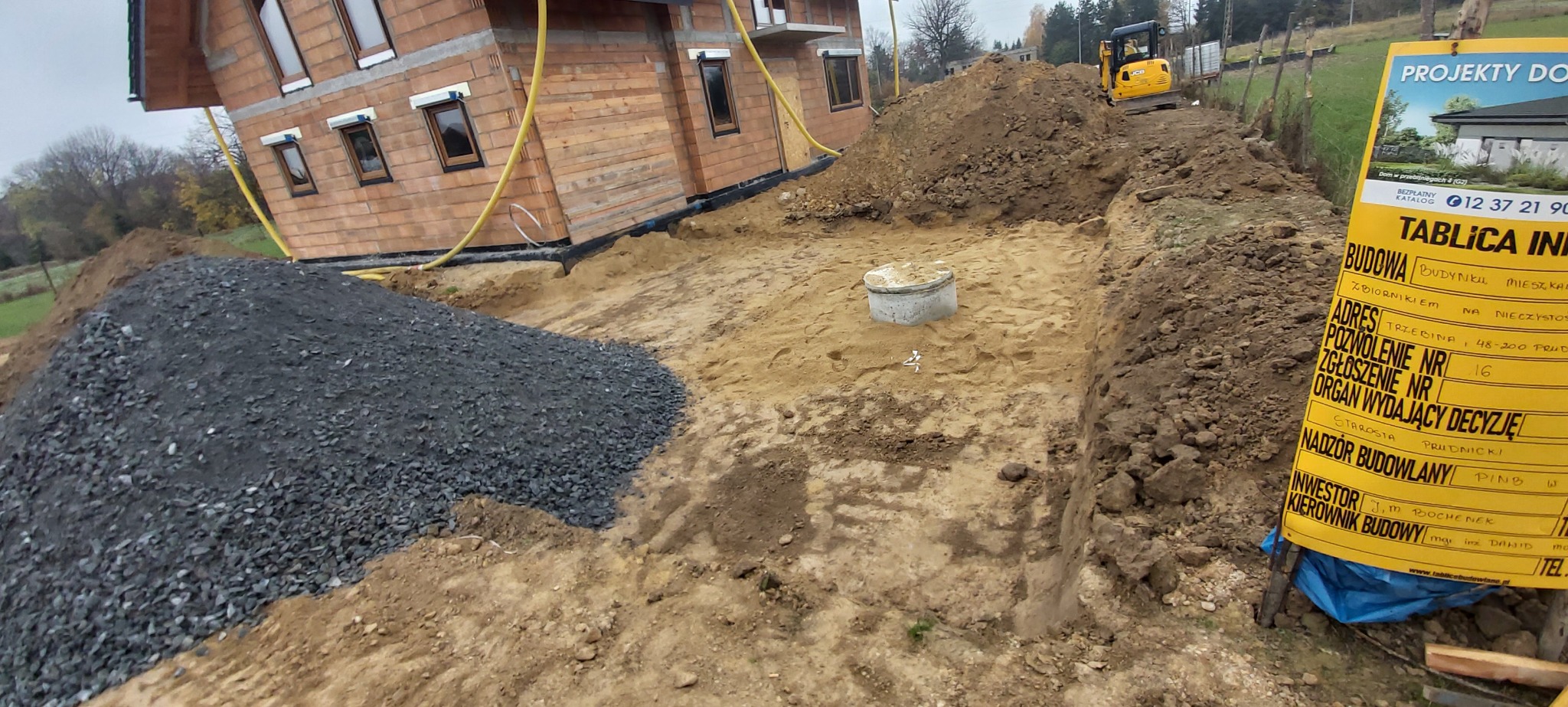 Wykop pod fundamenty domu w trakcie budowy, widoczny żółty mini-koparka, sterta grysu, betonowy zbiornik na szambo oraz tablica informacyjna budowy z danymi inwestora i kierownika budowy.