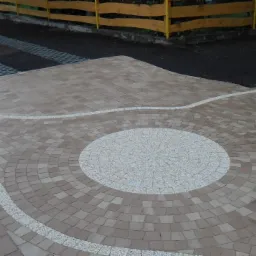 Ułożona kostka brukowa w odcieniach beżu z białymi akcentami tworzącymi wzór koła i łuków, widoczna część podjazdu z ażurowymi płytami, żółte drewniane ogrodzenie w tle.