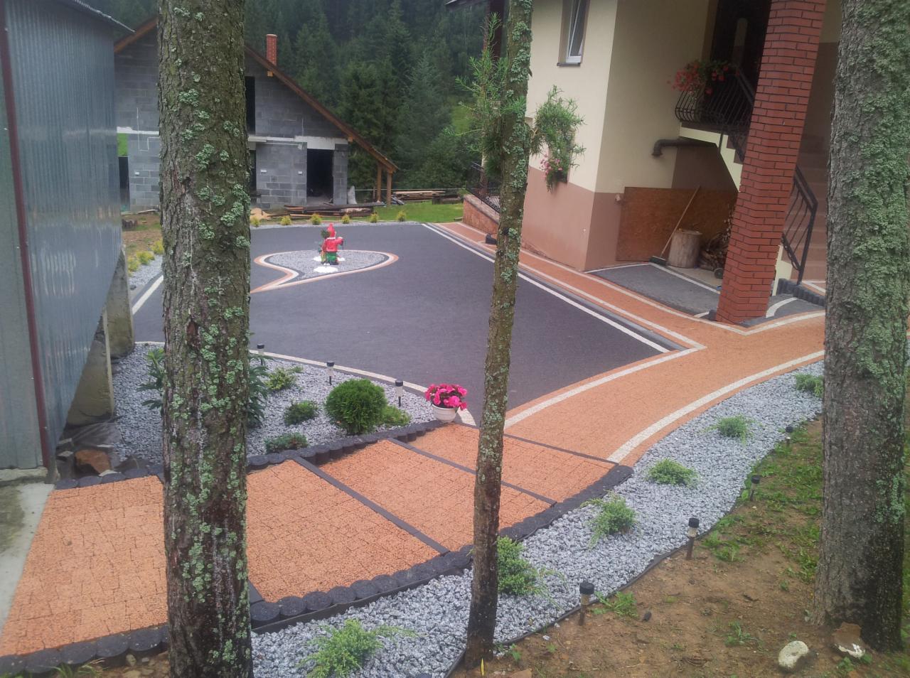 Ukończona aranżacja podwórka z ciemną nawierzchnią, jasną ścieżką z obrzeżami i szarym żwirem, widok zza pni drzew, w tle niedokończony budynek.